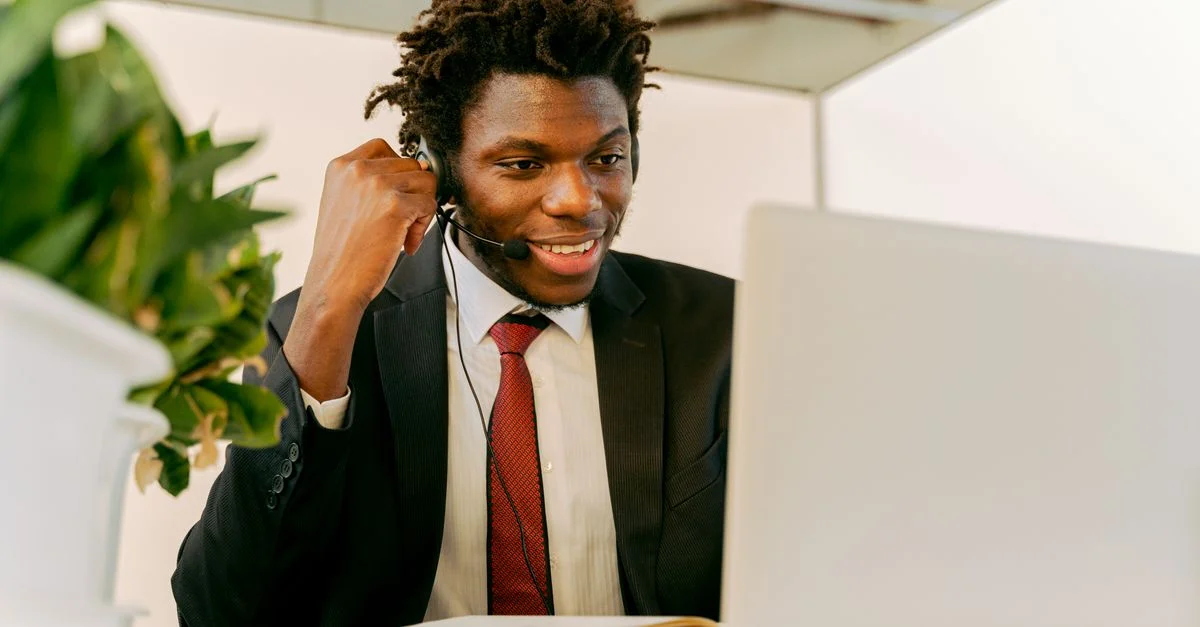 Glücklicher Geschäftsmann mit Afro-Haar, der ein Headset und einen Anzug trägt, während er in einem Büro an einem Laptop arbeitet.